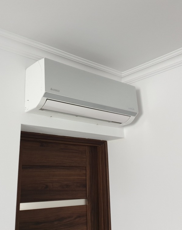 Klimatyzator ścienny marki Aircool zamontowany nad ciemnobrązowymi drzwiami z poziomymi panelami, widoczne białe listwy sufitowe.