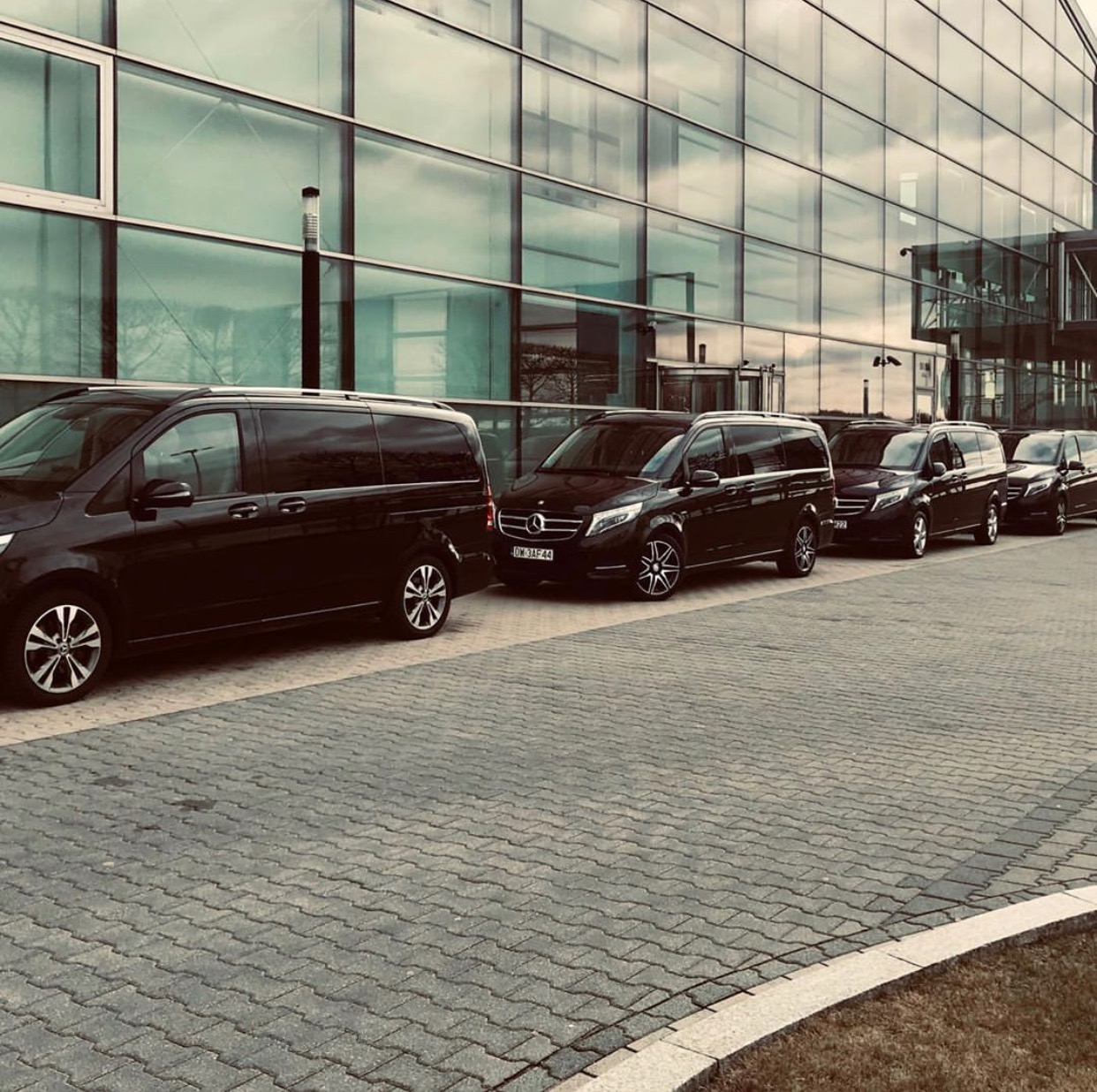 Flota czarnych busów Mercedes V-klasa ustawiona w rzędzie przed nowoczesnym szklanym budynkiem biurowym.