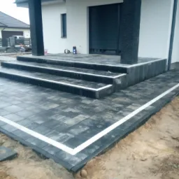 Wykończone schody wejściowe do domu z ciemnoszarej kostki brukowej z białym obramowaniem, widoczne czarne kolumny i elewacja domu w tle.