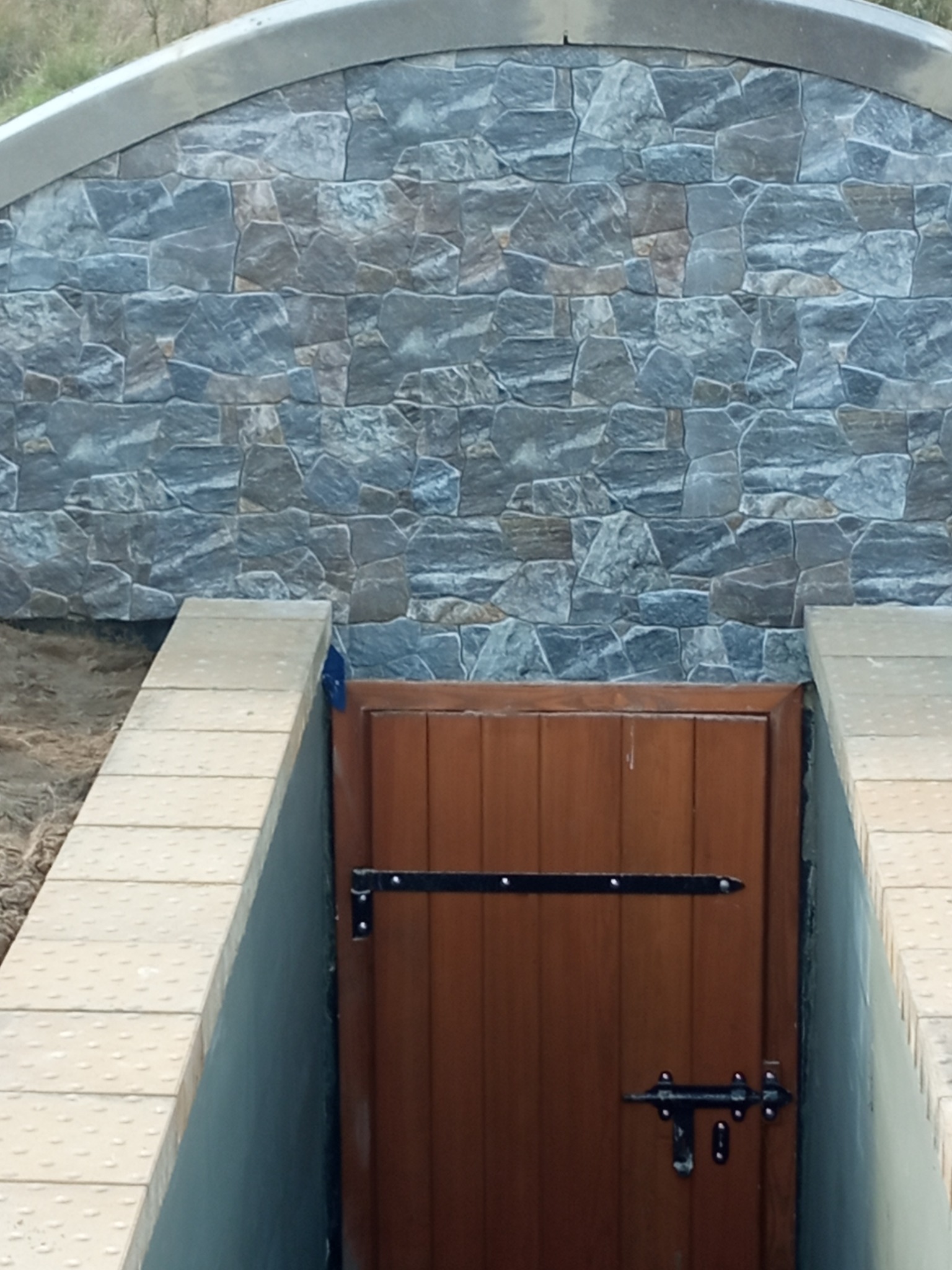 Wejście do piwnicy z drewnianymi drzwiami, wykończone kamienną elewacją i obramowane kostką brukową. Metalowe okucia drzwi.