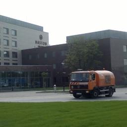 Sprzątanie Maszynowe Gąsior - Pomarańczowa zamiatarka Mercedes-Benz na tle nowoczesnego budynku hotelowego z zielonym trawnikiem z przodu.