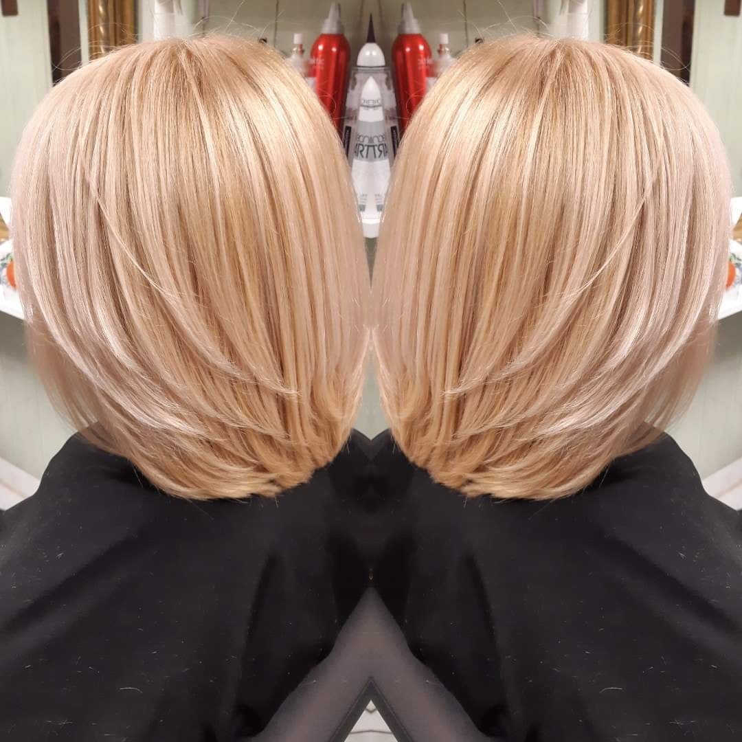 Blond włosy ścięte w warstwy, z widocznymi refleksami, prezentowane na tle półek z kosmetykami w salonie.