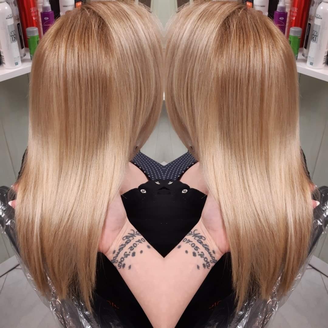 Blond włosy po koloryzacji z widocznymi odrostami, proste, sięgające do ramion, z delikatnym ombre, na tle półki z kosmetykami, z widocznymi tatuażami na nadgarstkach.