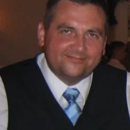 Portret mężczyzny w czarnej kamizelce i błękitnym krawacie, uśmiechającego się do kamery, na rozmytym tle wnętrza.