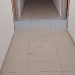 Beżowe płytki podłogowe w korytarzu, widoczne ślady po remoncie i fuga między płytkami. Ościeżnice drzwiowe z okleiną drewnopodobną po obu stronach korytarza.