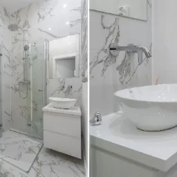 Nowoczesna łazienka z marmurowymi płytkami, kabiną prysznicową ze szklanymi drzwiami, białą umywalką nablatową na szafce z szufladami, chromowaną armaturą i dekoracyjną figurką ptaszka.