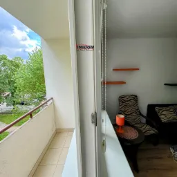 Widok z balkonu na zewnątrz, z fragmentem wnętrza mieszkania z pomarańczową lampką na stoliku i krzesłami w tle.