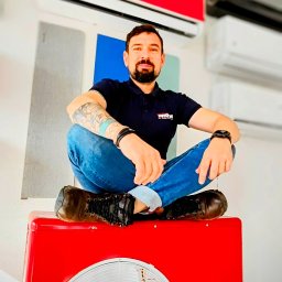 INTDOM MAREK KALINOWSKI - Mężczyzna z tatuażami siedzi po turecku na czerwonym wentylatorze, w tle wiszą klimatyzatory różnych kolorów i odcieni.