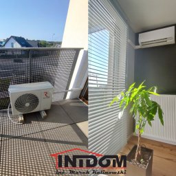 INTDOM MAREK KALINOWSKI - Klimatyzator Rotenso na balkonie z ażurową balustradą i klimatyzator w pomieszczeniu z żaluzjami, grzejnikiem i rośliną doniczkową. Kontrastowe ujęcie.