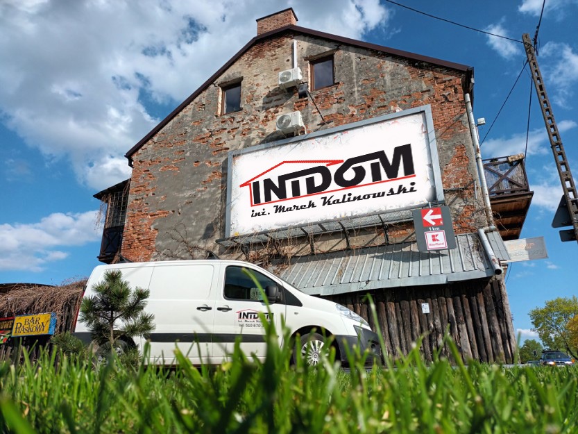 Budynek z zamontowanymi klimatyzatorami, na którym wisi baner reklamowy firmy, na pierwszym planie trawa i samochód dostawczy z logo firmy.