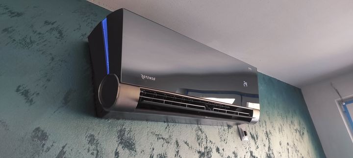 Czarny klimatyzator Rotenso z wyświetlaczem temperatury 24 stopnie Celsjusza, zamontowany na zielono-niebieskiej ścianie o nieregularnej fakturze, widoczny fragment okna