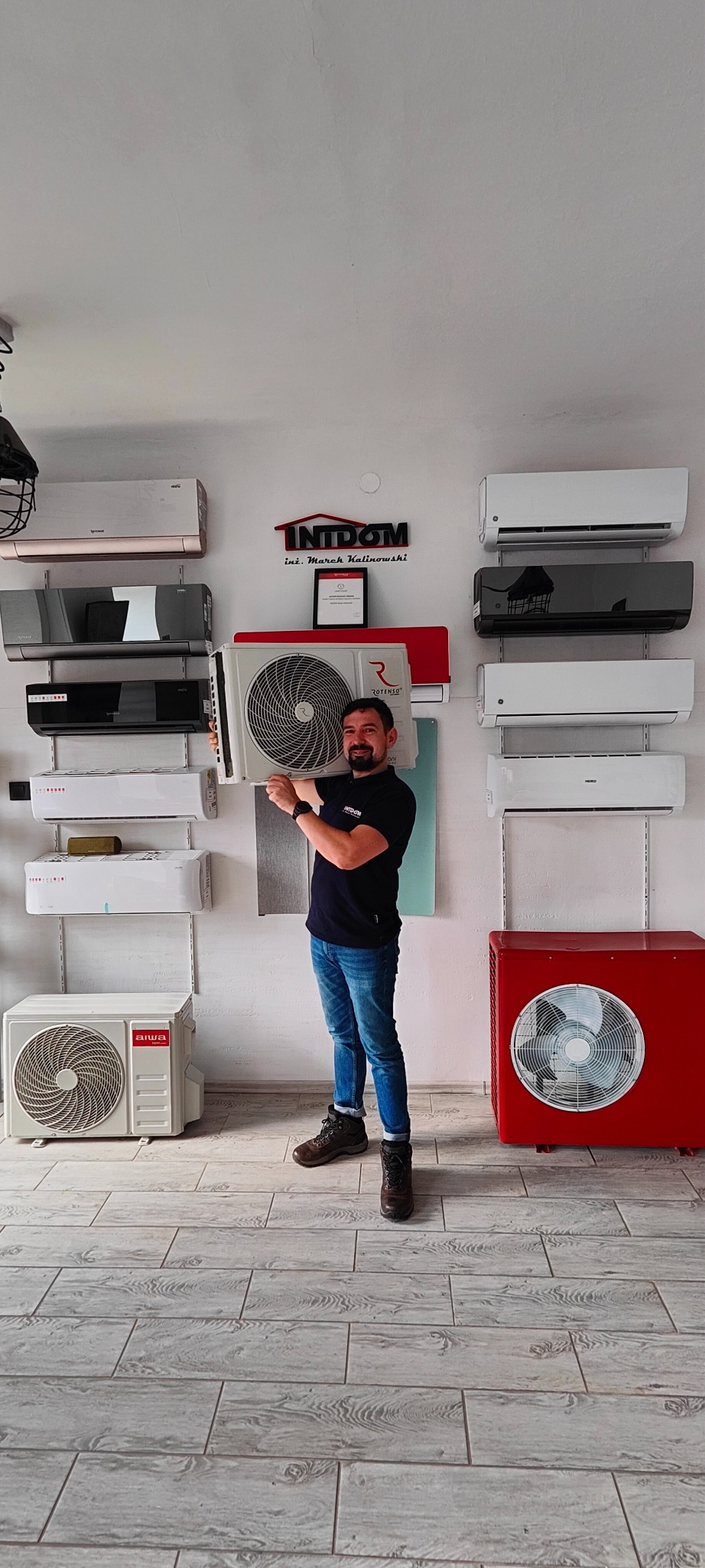 Mężczyzna w czarnej koszulce i jeansach prezentuje model klimatyzatora Rotenso na tle ściany z ekspozycją różnych urządzeń klimatyzacyjnych i logo firmy Intidom.