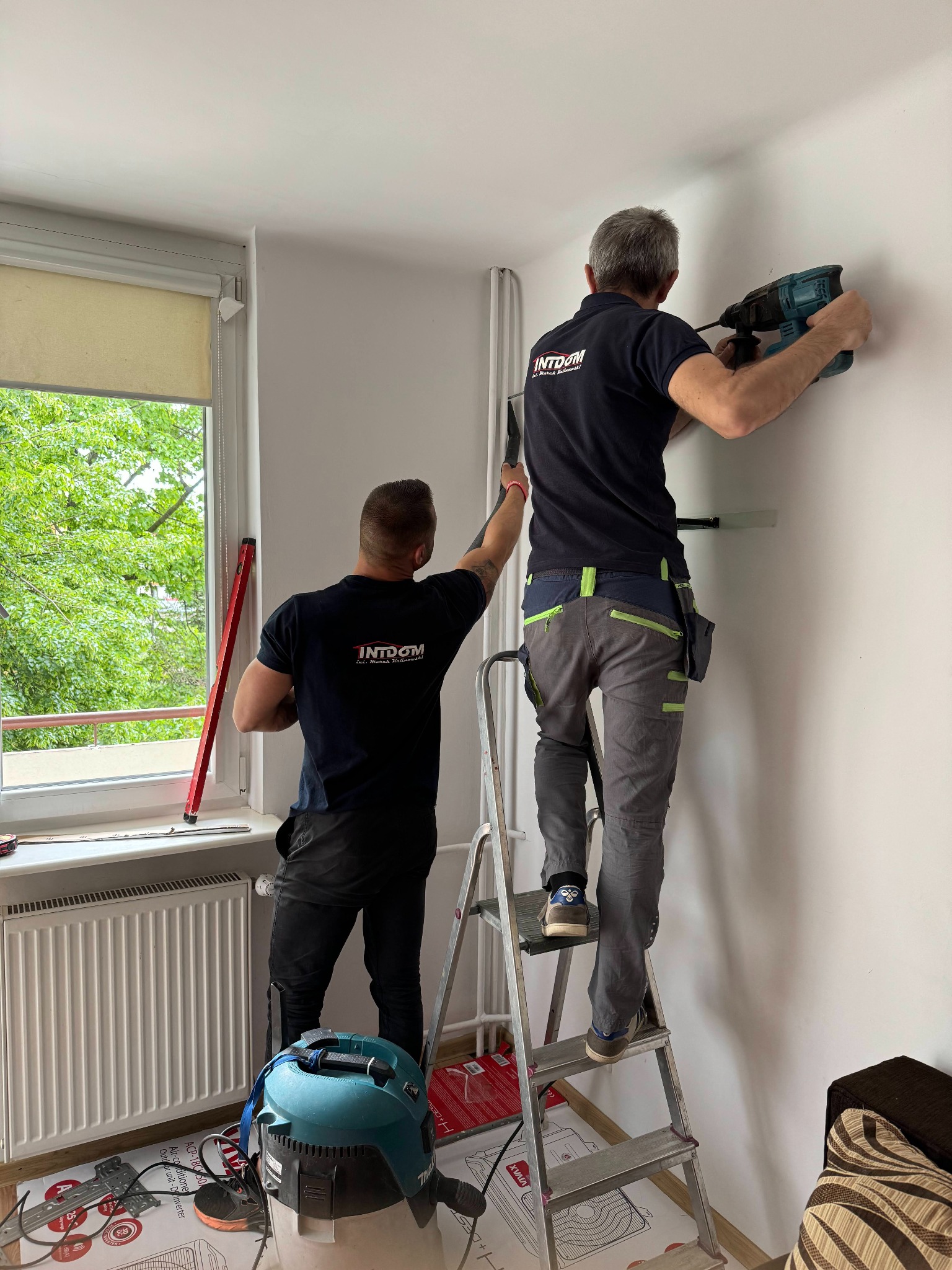 Dwóch monterów w trakcie instalacji urządzenia na ścianie wewnątrz pomieszczenia. Jeden wierci wiertarką udarową, stojąc na aluminiowej drabinie, drugi przy oknie z poziomicą w dłoni. Na podłodze...