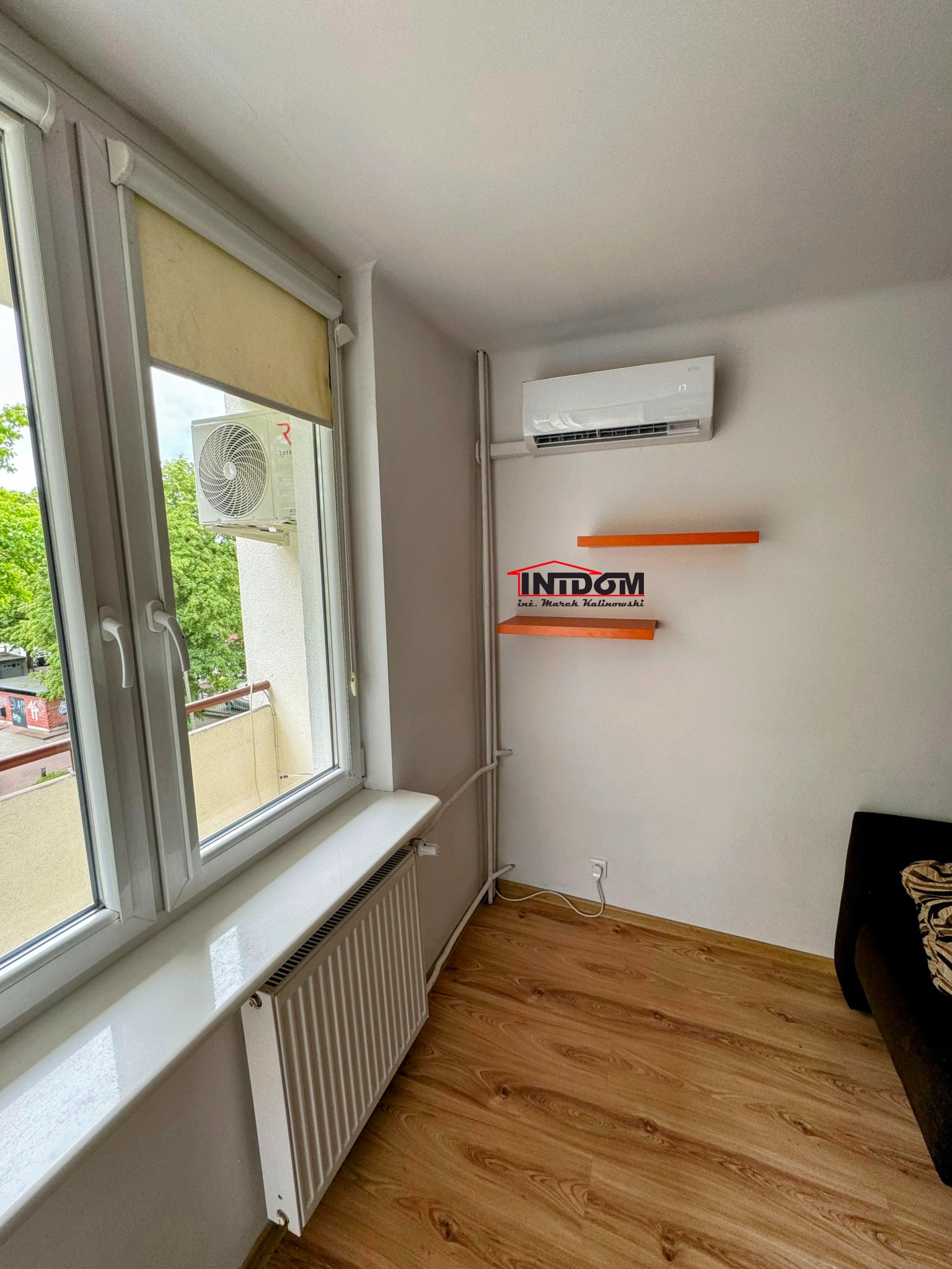 Wnętrze pokoju w Puławach z zamontowaną klimatyzacją ścienną, widoczną przez okno jednostką zewnętrzną na balkonie oraz dwoma pomarańczowymi półkami na ścianie.
