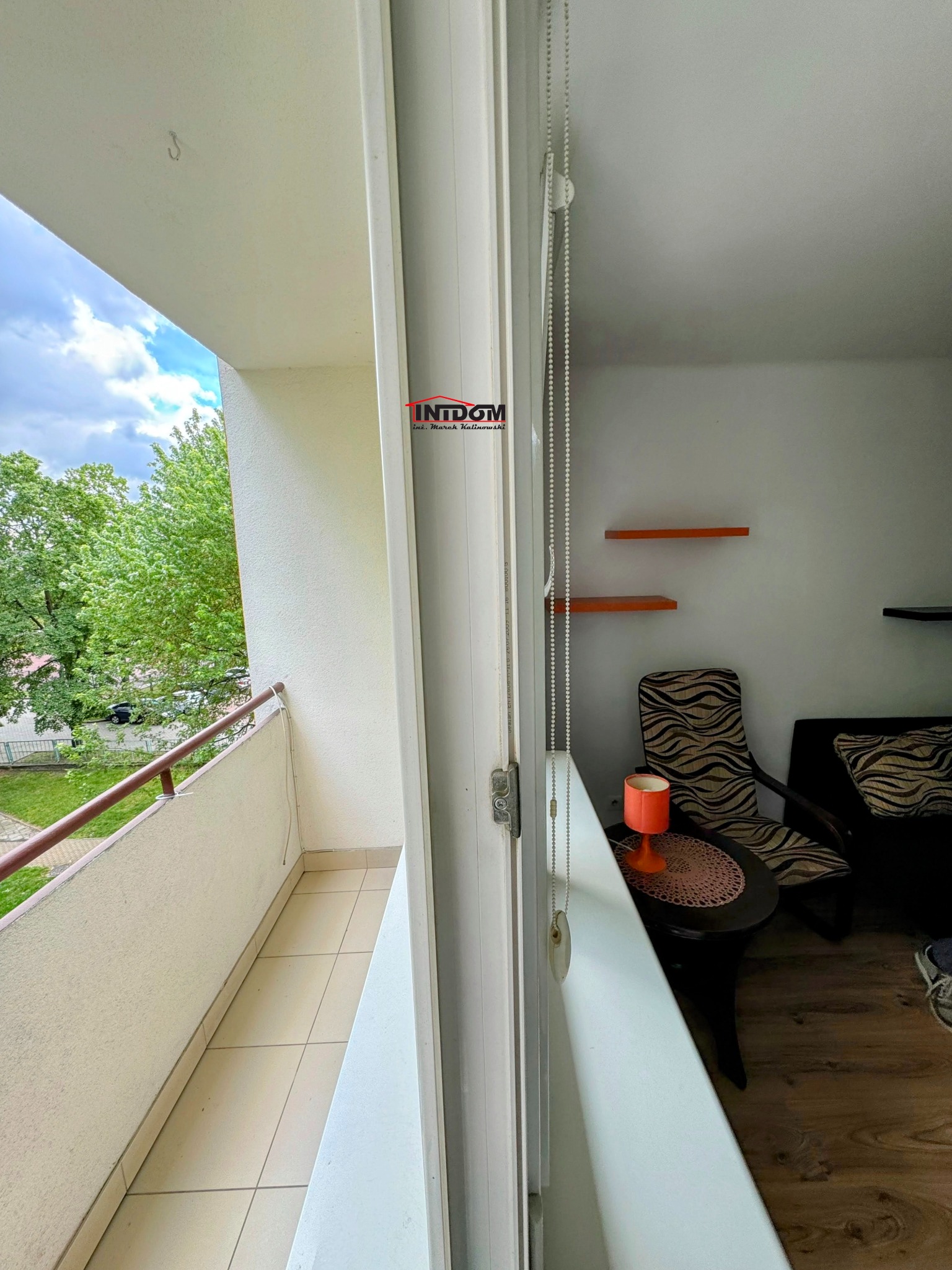 Widok z balkonu na zewnątrz, z fragmentem wnętrza mieszkania z pomarańczową lampką na stoliku i krzesłami w tle.