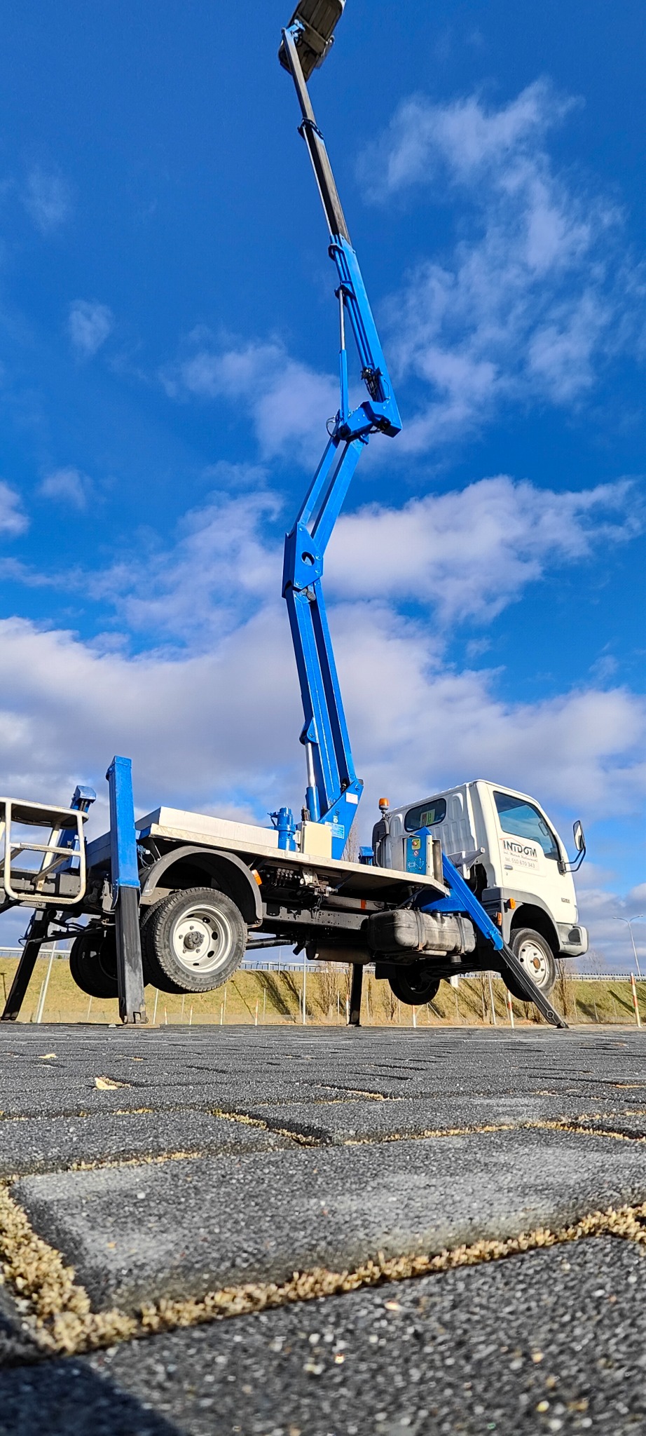Podnośnik koszowy na białym samochodzie marki Indom z wysięgnikiem w kolorze niebieskim, ustawiony na kostce brukowej, z błękitnym niebem i chmurami w tle.