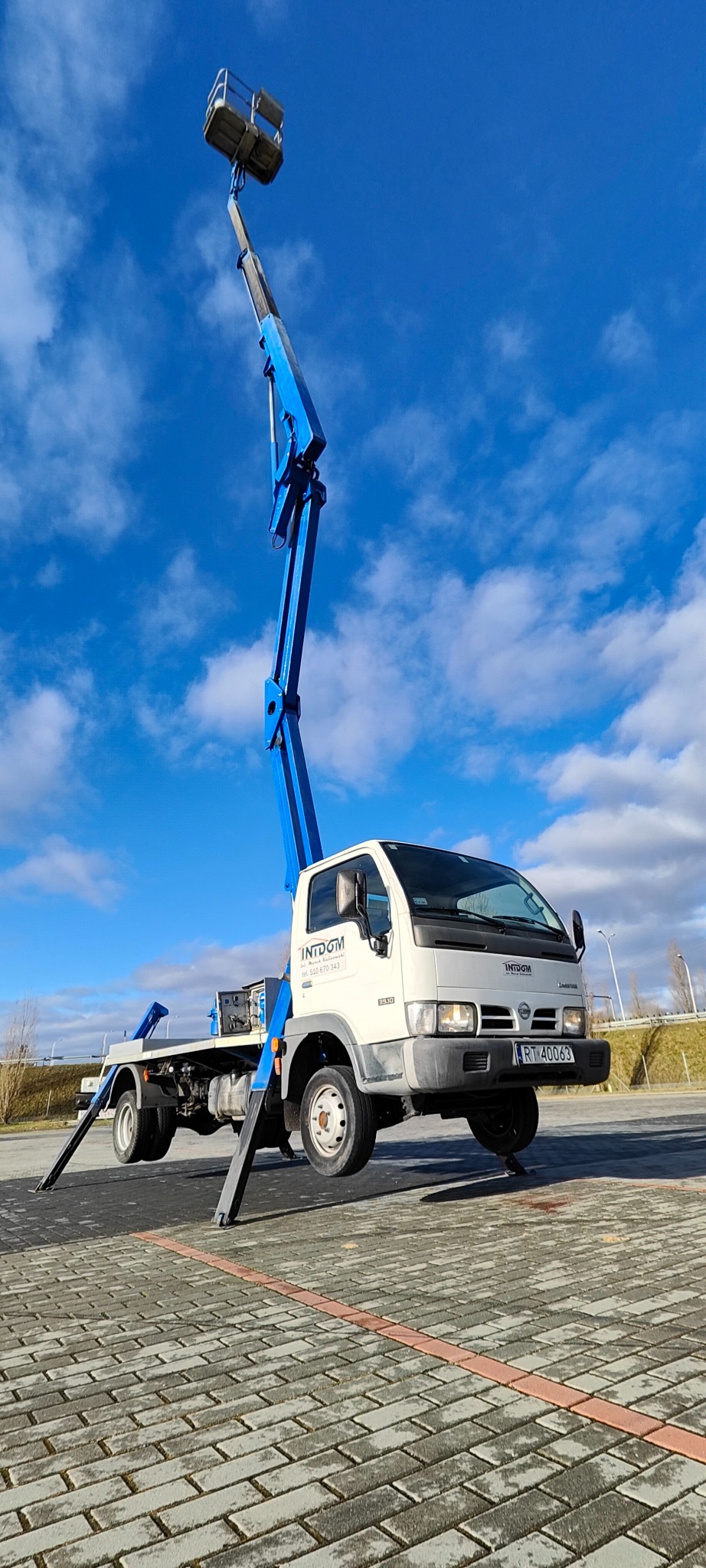 Biała ciężarówka Nissan z niebieskim podnośnikiem koszowym na parkingu z kostki brukowej, błękitne niebo w tle.