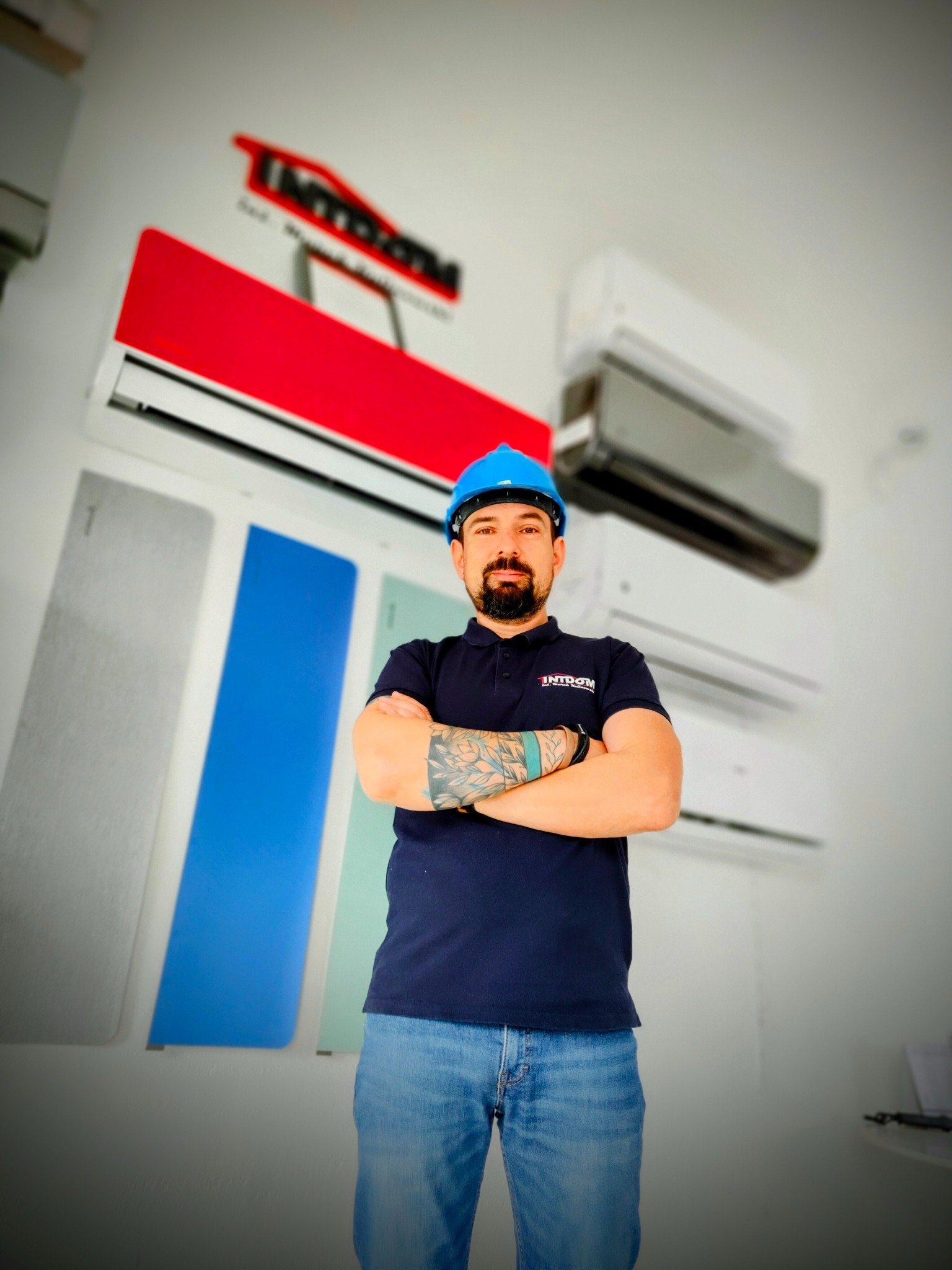 Mężczyzna w niebieskim kasku i koszulce z logo firmy na tle ściany z zamontowanymi klimatyzatorami o różnych kolorach obudowy.