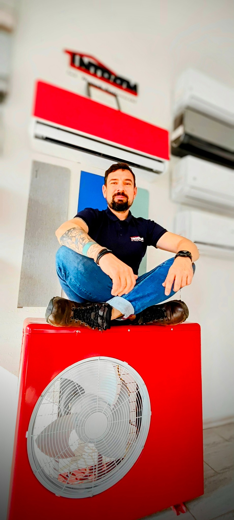 Mężczyzna z tatuażami siedzi po turecku na czerwonym wentylatorze, w tle wiszą klimatyzatory różnych kolorów i odcieni.