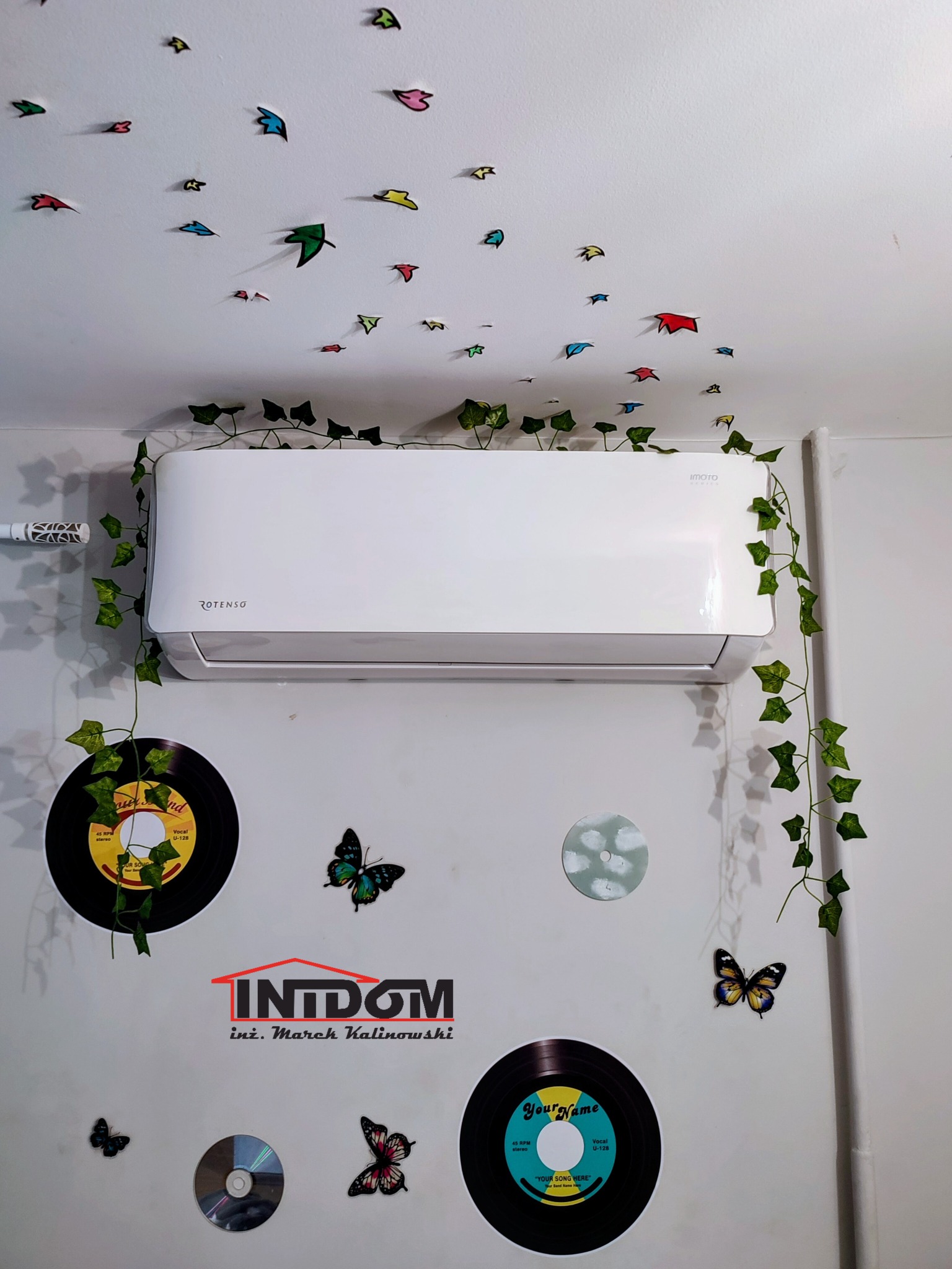 Biała klimatyzacja Rotenso w pokoju z dekoracjami ściennymi, montaż w Puławach. Rośliny pnące i naklejki motyli dodają uroku. Widoczne płyty winylowe i logo firmy.