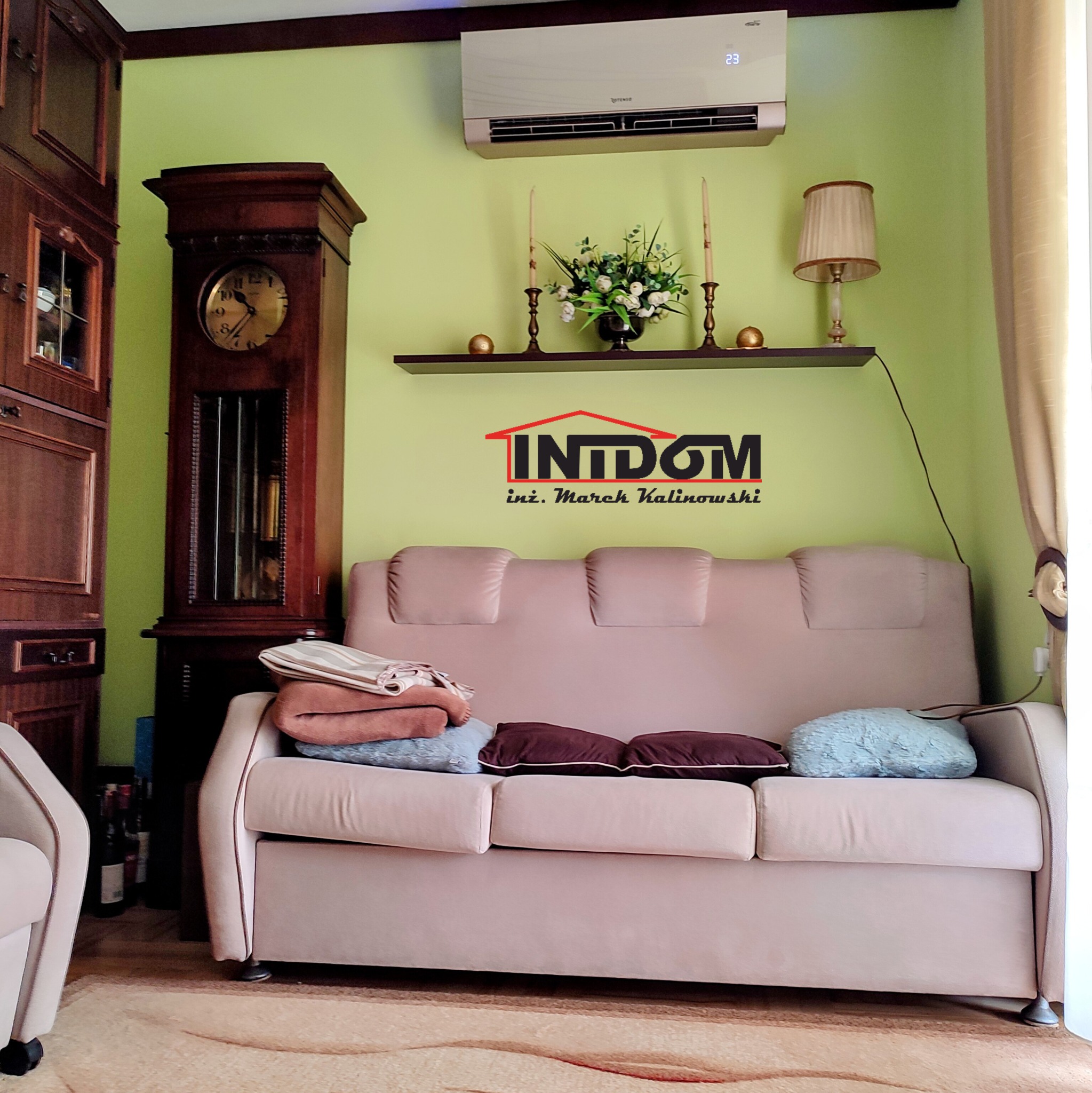 Klimatyzator Rotenso zamontowany nad sofą w salonie z zielonymi ścianami, obok stoi zegar dziadka i witryna. Na ścianie półka z dekoracjami.