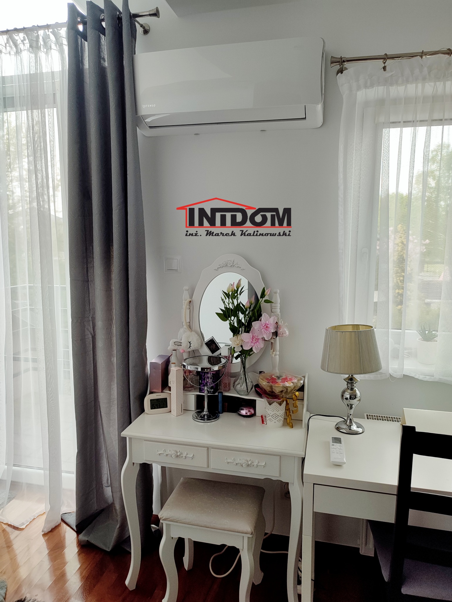 Biały klimatyzator Rotenso nad toaletką w pokoju z oknami i zasłonami. Widoczny pilot do klimatyzacji na stoliku obok. Logo firmy na ścianie.