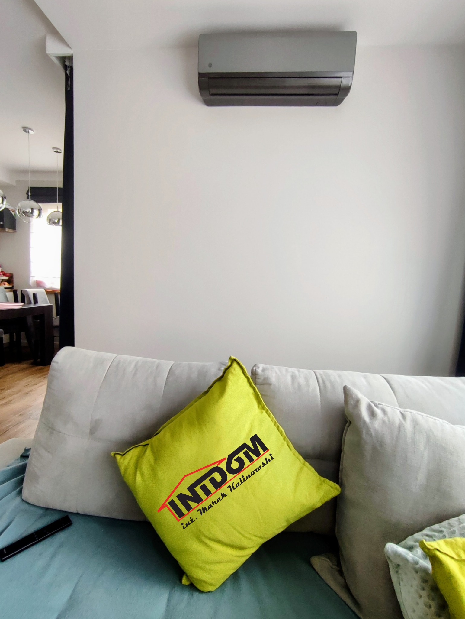 Szary klimatyzator ścienny w nowoczesnym salonie, widoczny zza sofy z zieloną poduszką z logo firmy. W tle fragment jadalni z lampami.