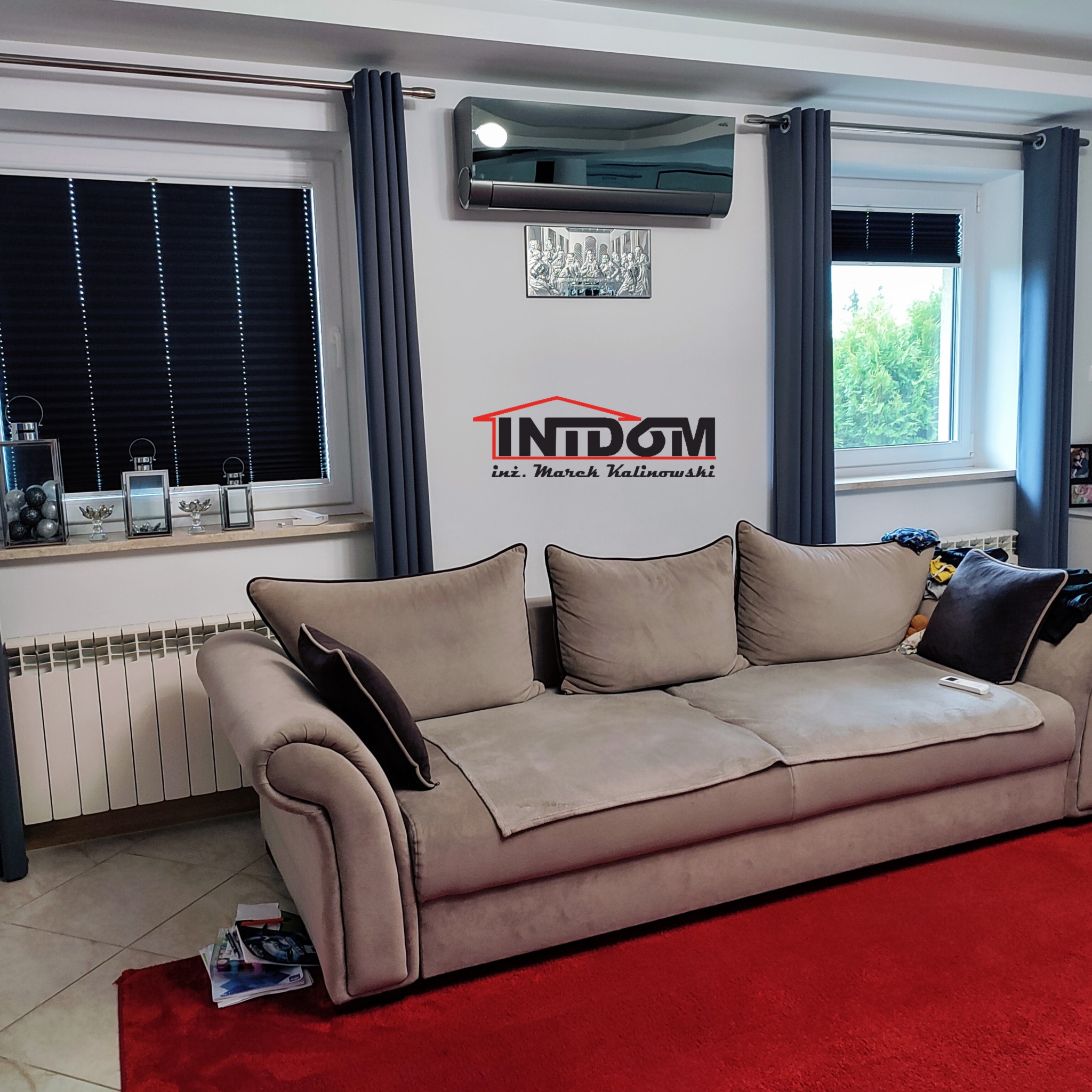 Nowoczesny klimatyzator ścienny w salonie z szarymi zasłonami i żaluzjami. Widoczny beżowy narożnik i czerwony dywan. Nad kanapą logo firmy.