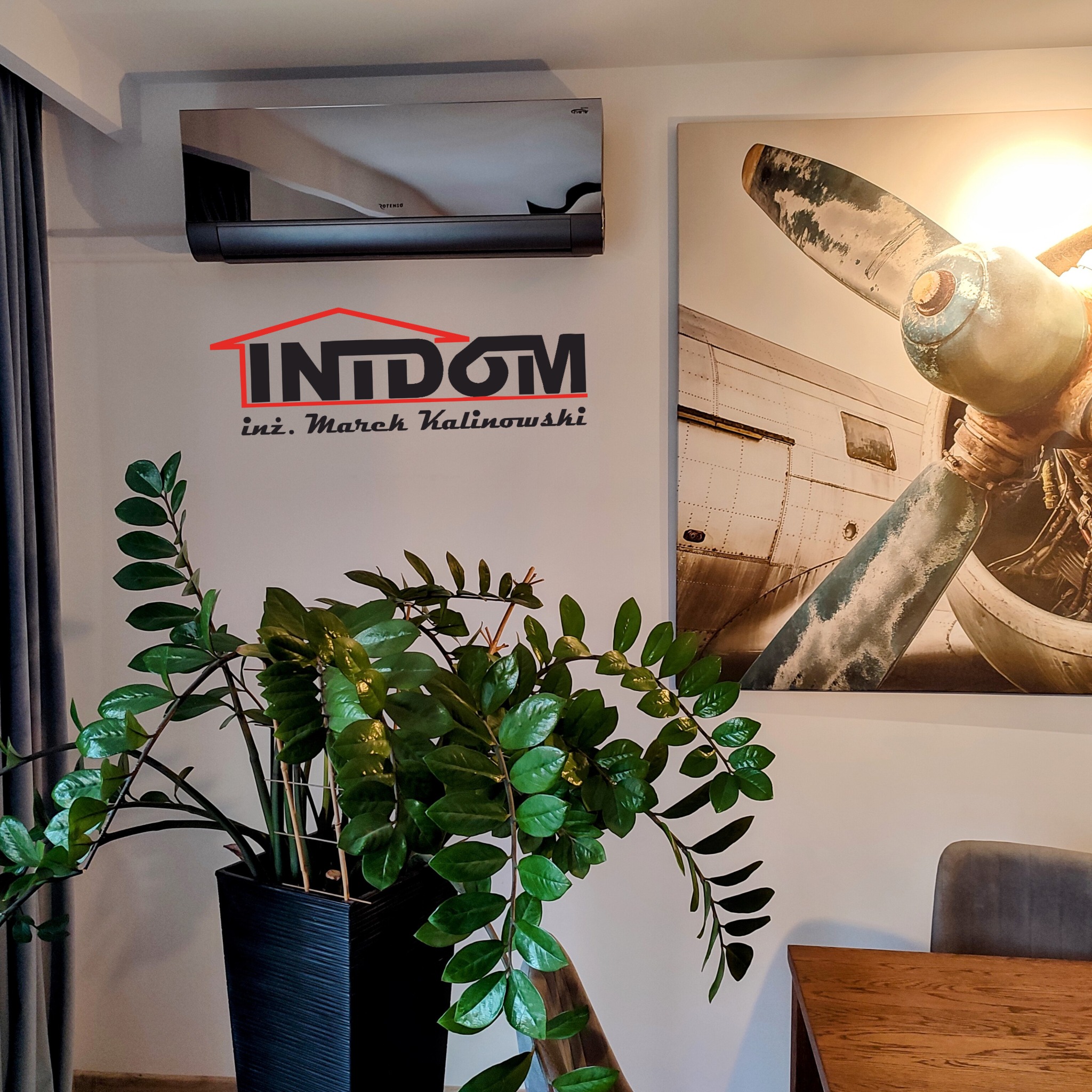 Nowoczesny klimatyzator Rotenso w salonie z rośliną i obrazem samolotu. Czarny model na białej ścianie, widoczne logo firmy instalacyjnej.