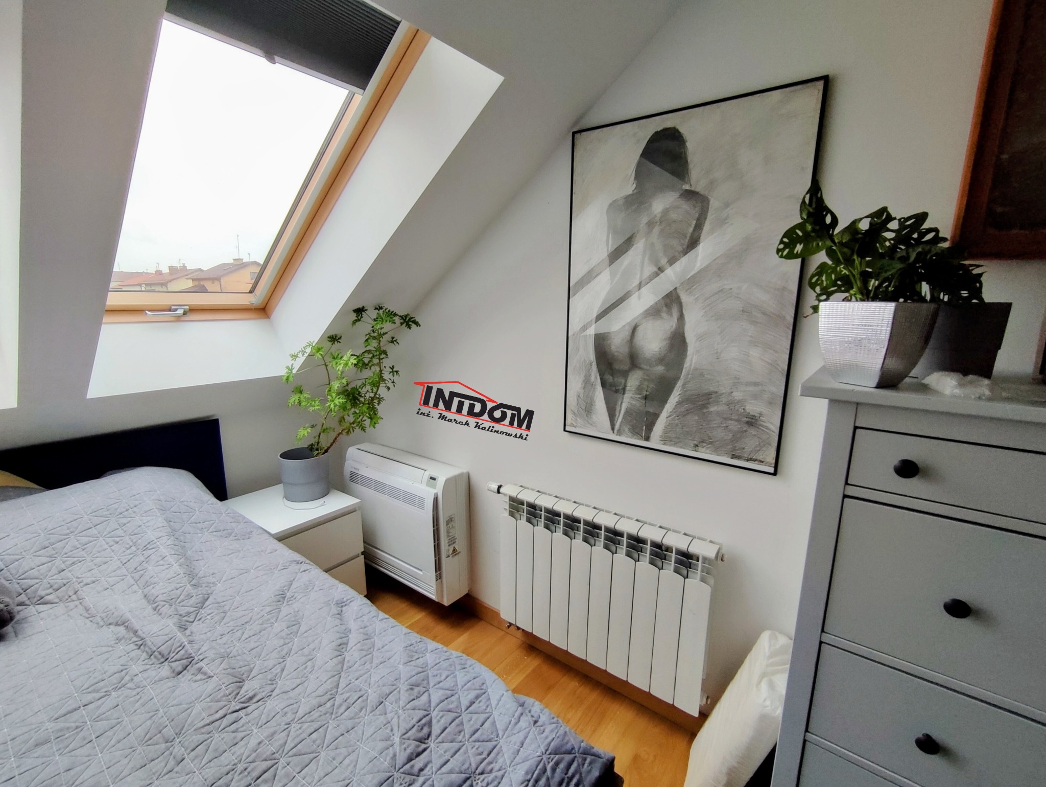 Pokój na poddaszu z grzejnikiem i klimatyzatorem obok łóżka. Na ścianie obraz i logo firmy. Okno dachowe wpuszcza światło.