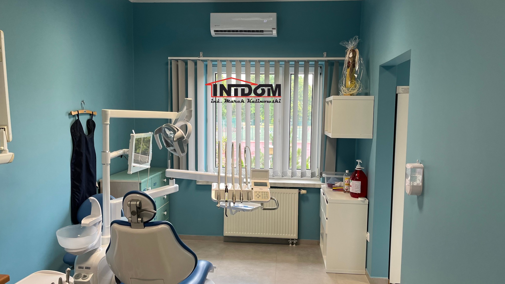 Jasne wnętrze gabinetu dentystycznego z klimatyzatorem nad oknem z żaluzjami pionowymi. Widoczne krzesło dentystyczne i sprzęt medyczny.