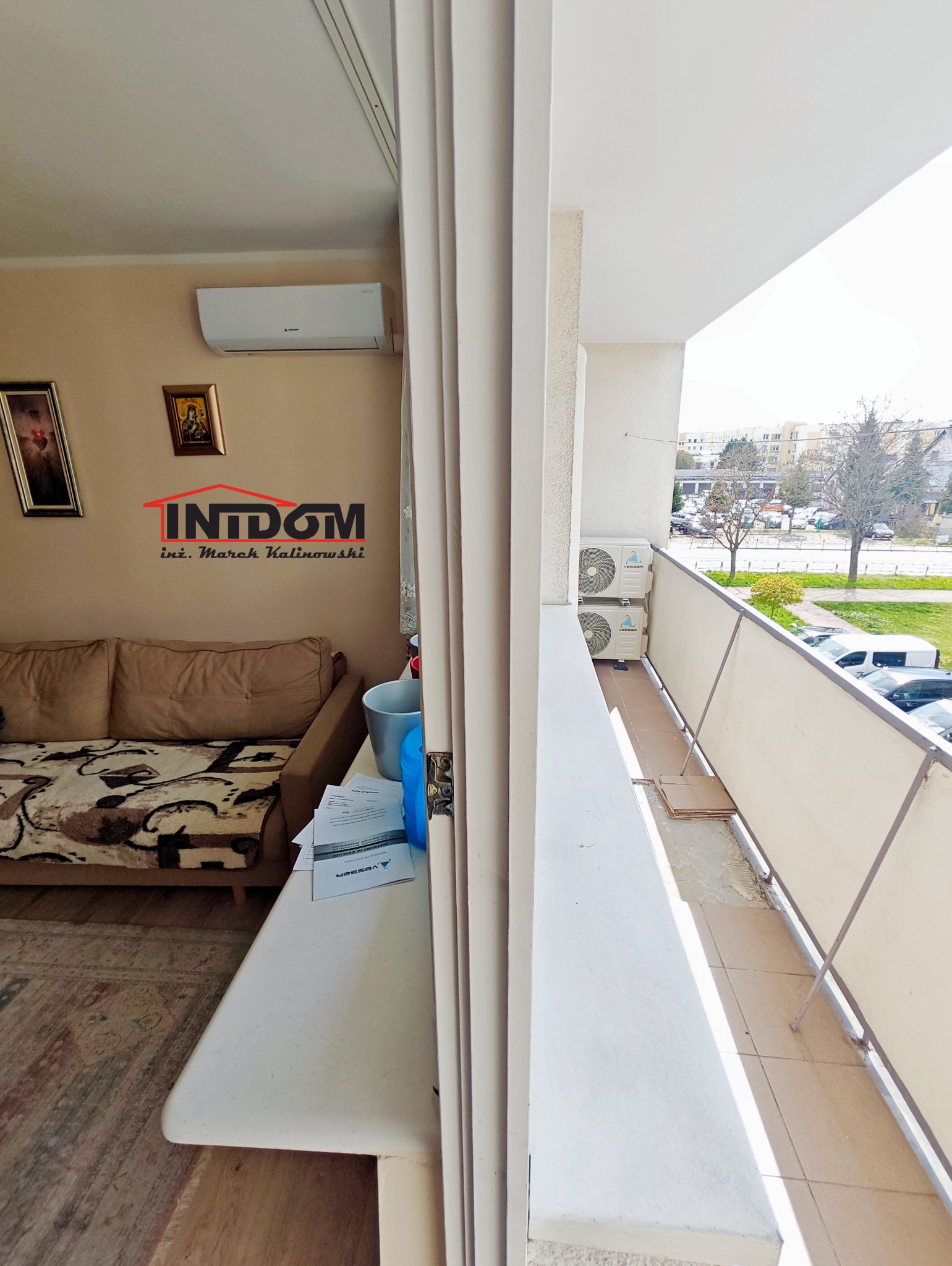 Widok z balkonu na dwa zamontowane klimatyzatory na zewnątrz. W tle budynki mieszkalne i zieleń. Wnętrze mieszkania z klimatyzatorem widoczne po lewej stronie.