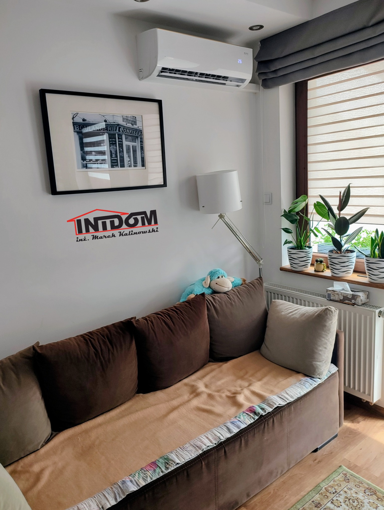 Biały klimatyzator ścienny w salonie nad sofą, z widocznym wyświetlaczem temperatury. Na ścianie grafika w czarnej ramie i logo firmy. Stonowana kolorystyka wnętrza.