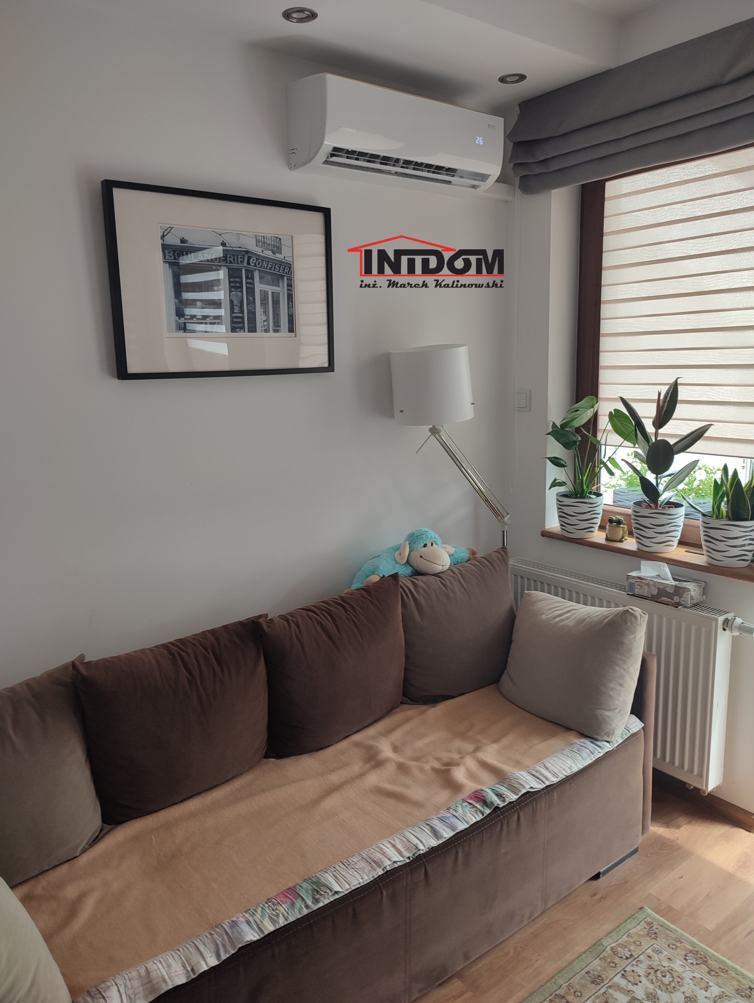 Biały klimatyzator ścienny w salonie nad brązową sofą, obok okna z roletami rzymskimi i roślinami doniczkowymi. Na ścianie czarno-biała grafika.