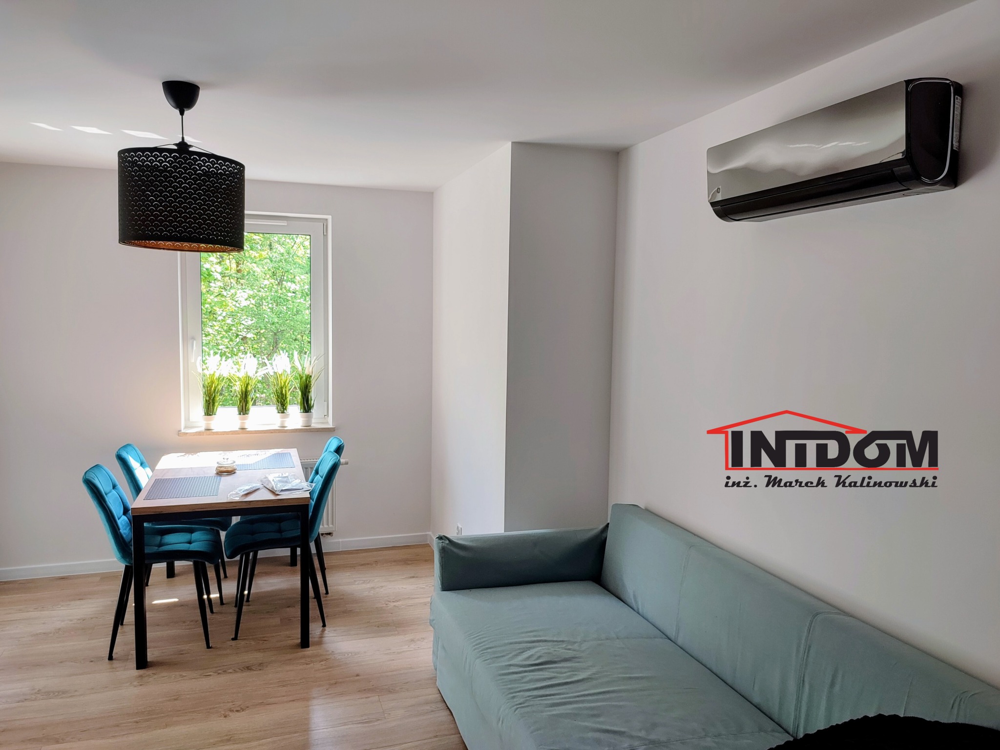 Nowoczesny, czarny klimatyzator ścienny w salonie z turkusowymi krzesłami i sofą. Widoczne logo firmy na ścianie. Jasne wnętrze z oknem i roślinami.