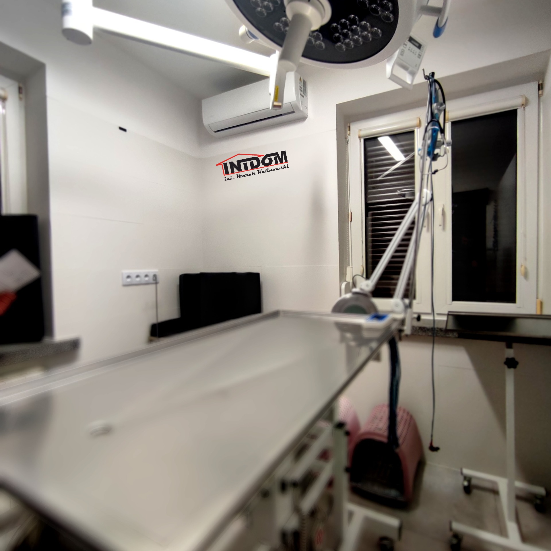 Jasne wnętrze gabinetu weterynaryjnego z klimatyzacją, stołem operacyjnym i lampą zabiegową. Widoczny logo firmy na ścianie.