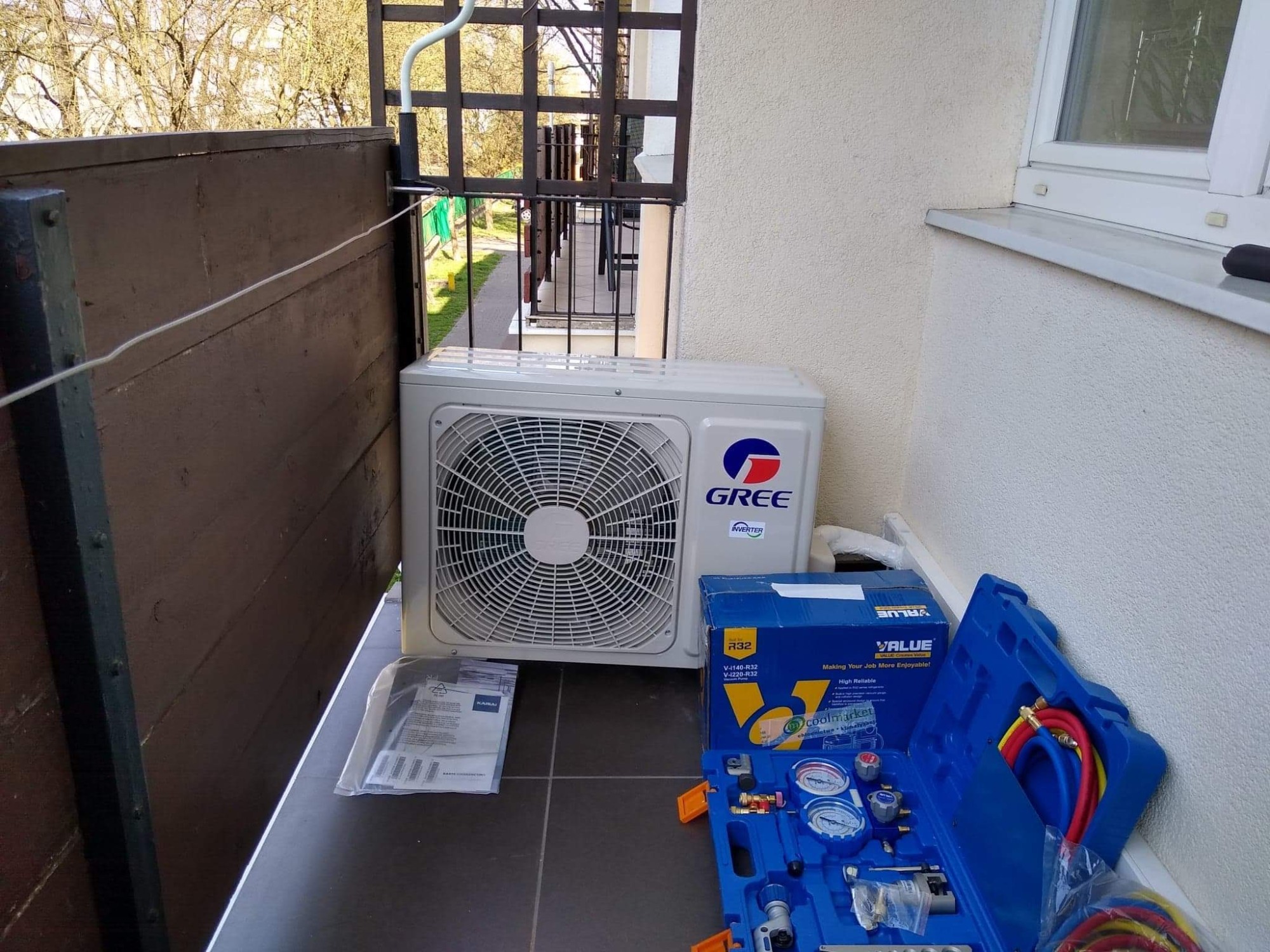 Jednostka zewnętrzna klimatyzatora GREE na balkonie, obok narzędzia w niebieskiej walizce i instrukcja montażu w folii.