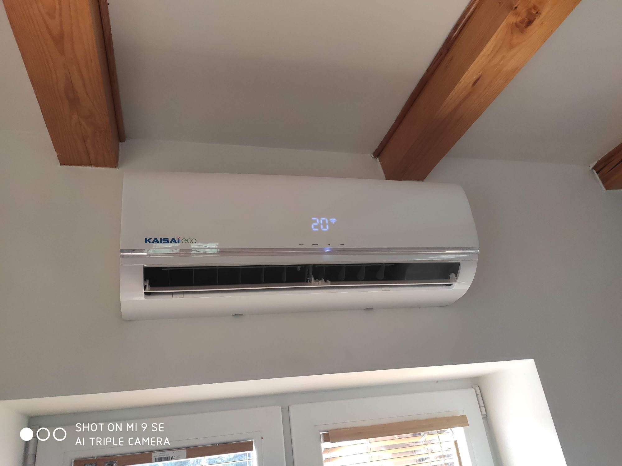 Biały klimatyzator Kaisai Eco zamontowany na ścianie w pomieszczeniu z drewnianymi belkami stropowymi, ustawiony na 20 stopni Celsjusza.