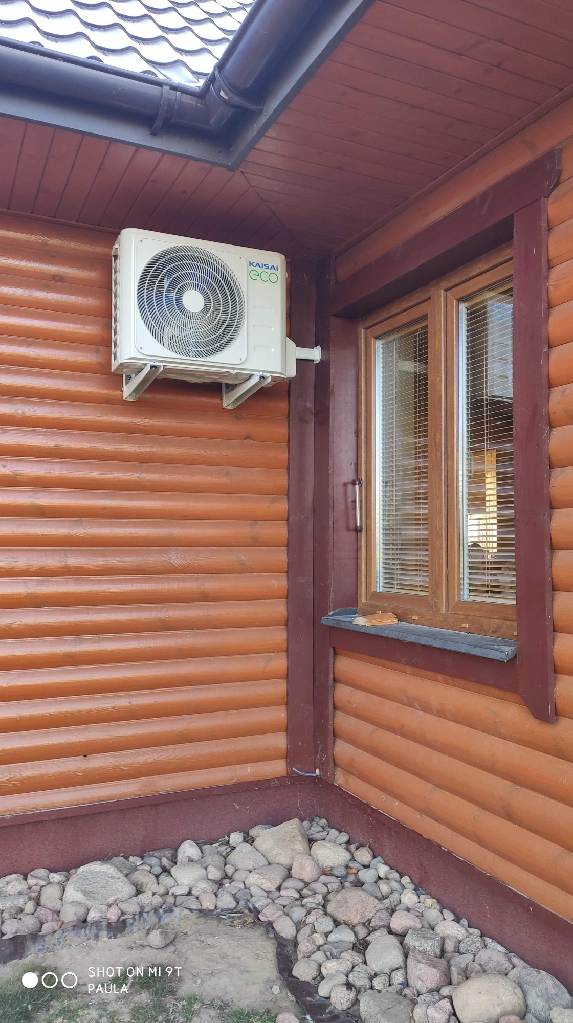 Biała jednostka klimatyzacji Kaisai ECO zamontowana na brązowej, drewnianej ścianie domu z widocznym oknem i kamiennym obłożeniem u podstawy.