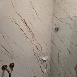 REMTOM - Fragment łazienki z białymi płytkami imitującymi marmur z brązowymi żyłami, widoczna bateria umywalkowa w kolorze miedzi, szklana kabina prysznicowa oraz gniazdka elektryczne.