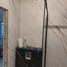 REMTOM - Wnętrze łazienki w trakcie prac wykończeniowych: częściowo położone płytki imitujące marmur z brązowymi żyłami, czarna zabudowa WC, widoczna konstrukcja kabiny prysznicowej, akcesoria i chemia...
