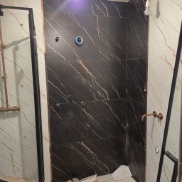 REMTOM - Łazienka w trakcie montażu płytek: ściana wyłożona czarnymi płytkami z białymi żyłami, obok ściana z białych płytek z brązowymi żyłami, widoczne otwory na instalacje, na podłodze zabezpieczona...