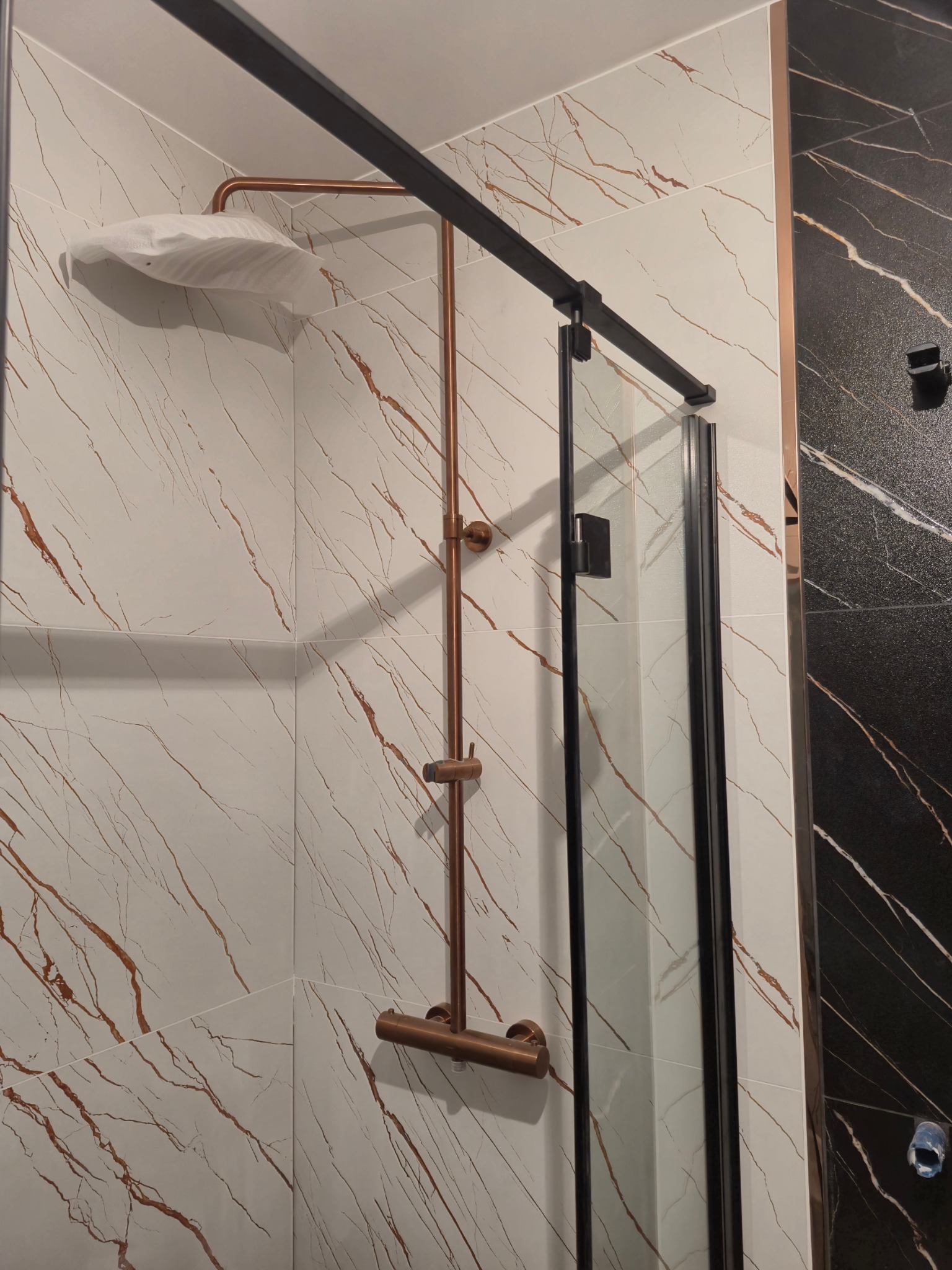 Nowoczesna kabina prysznicowa z miedzianą armaturą i czarnymi profilami, białe płytki z marmurkowym wzorem, głowica prysznicowa owinięta folią ochronną.