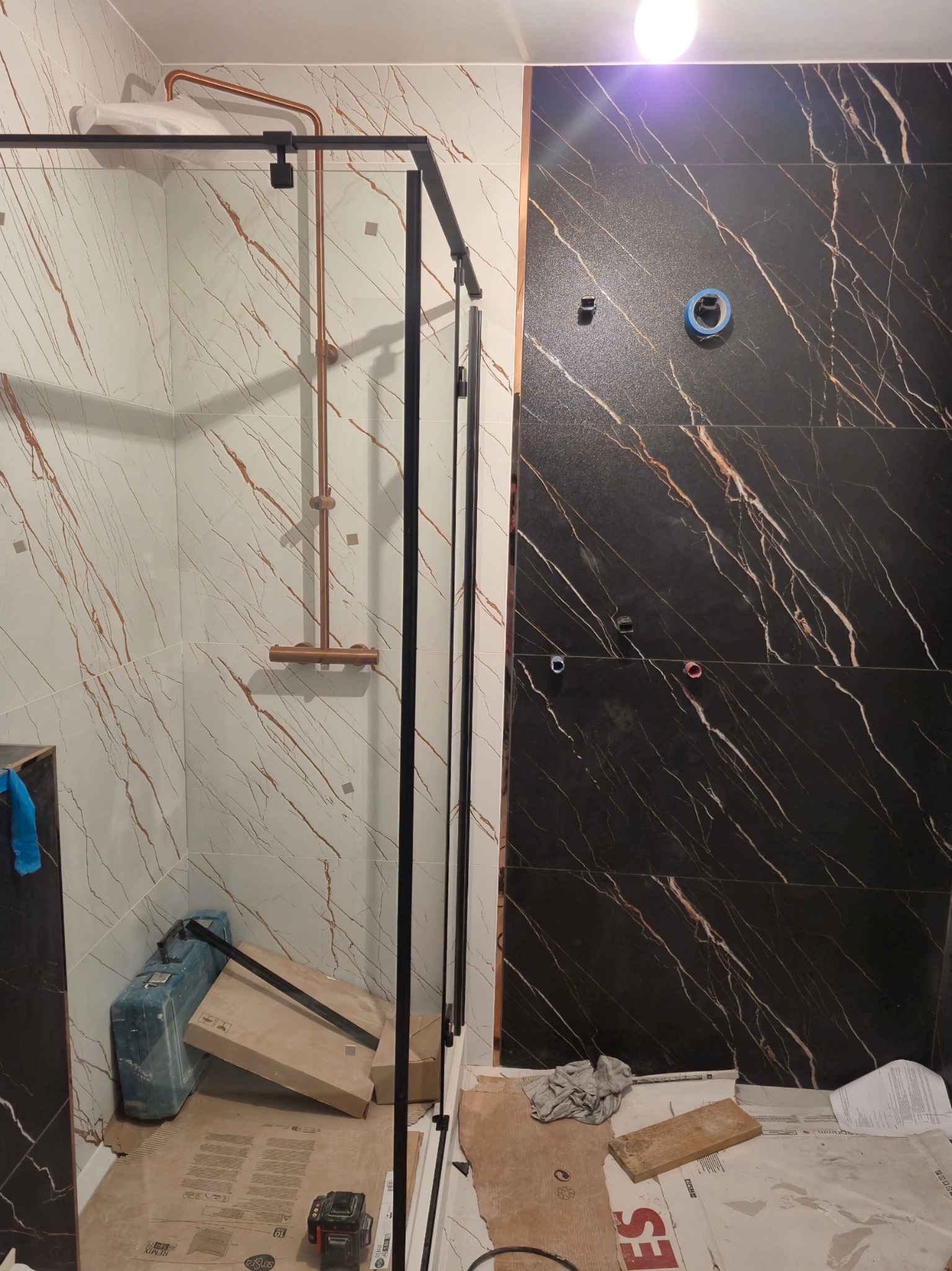 Montaż kabiny prysznicowej z czarnymi profilami na tle marmurowych płytek w odcieniach bieli i czerni, widoczne narzędzia i materiały budowlane na podłodze.