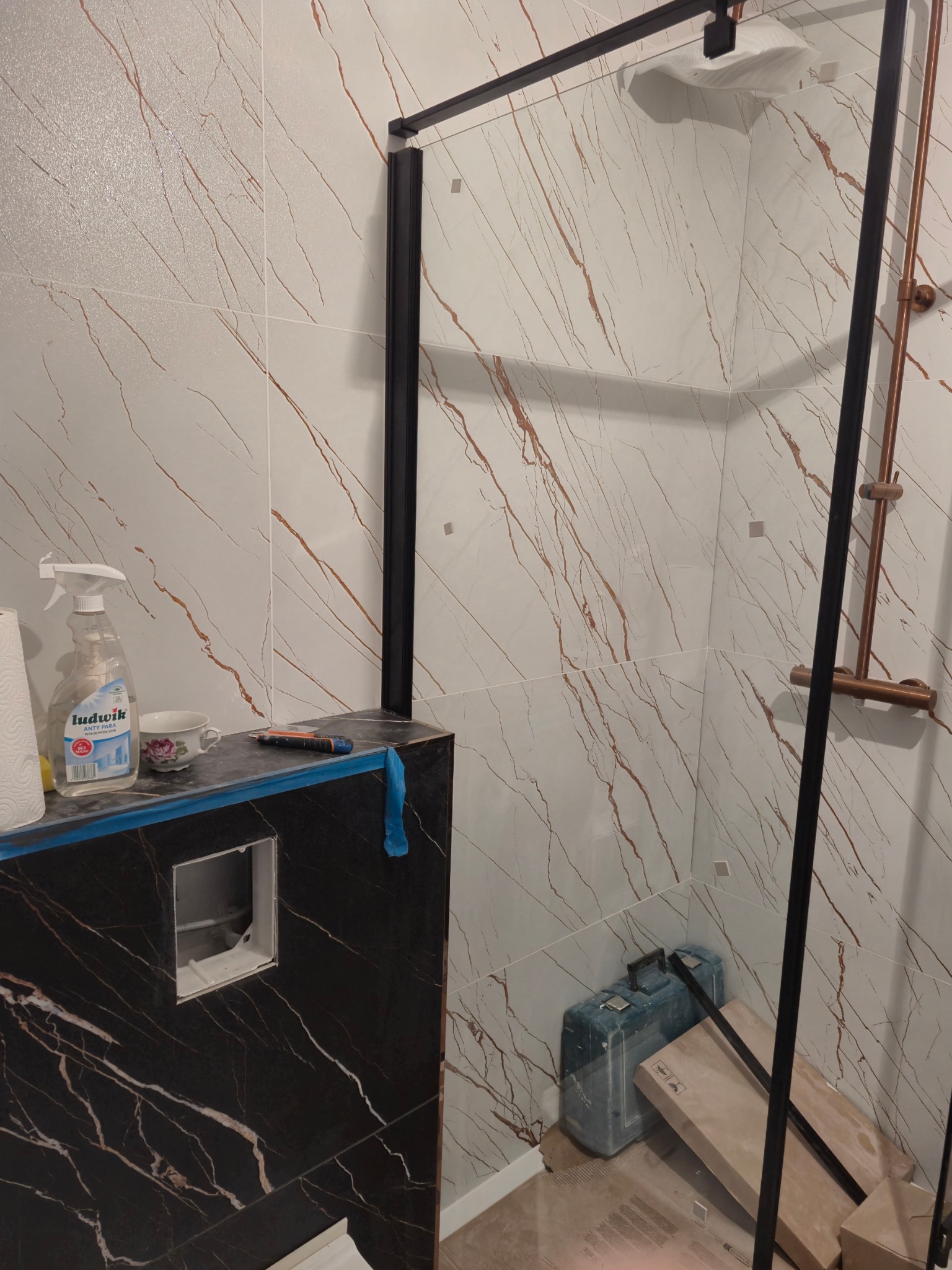 Niedokończona łazienka z kabiną prysznicową z czarną ramą, biało-brązowe płytki imitujące marmur na ścianach, czarne płytki na obudowie stelaża podtynkowego, widoczny środek czyszczący Ludwik...