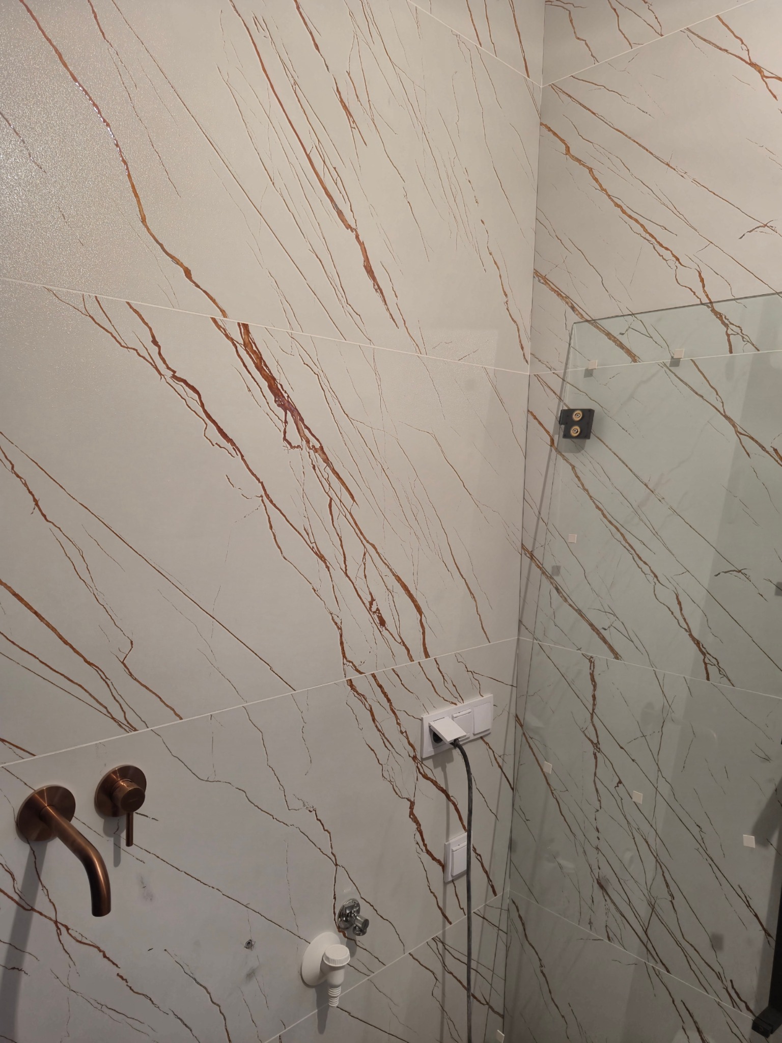 Fragment łazienki z białymi płytkami imitującymi marmur z brązowymi żyłami, widoczna bateria umywalkowa w kolorze miedzi, szklana kabina prysznicowa oraz gniazdka elektryczne.