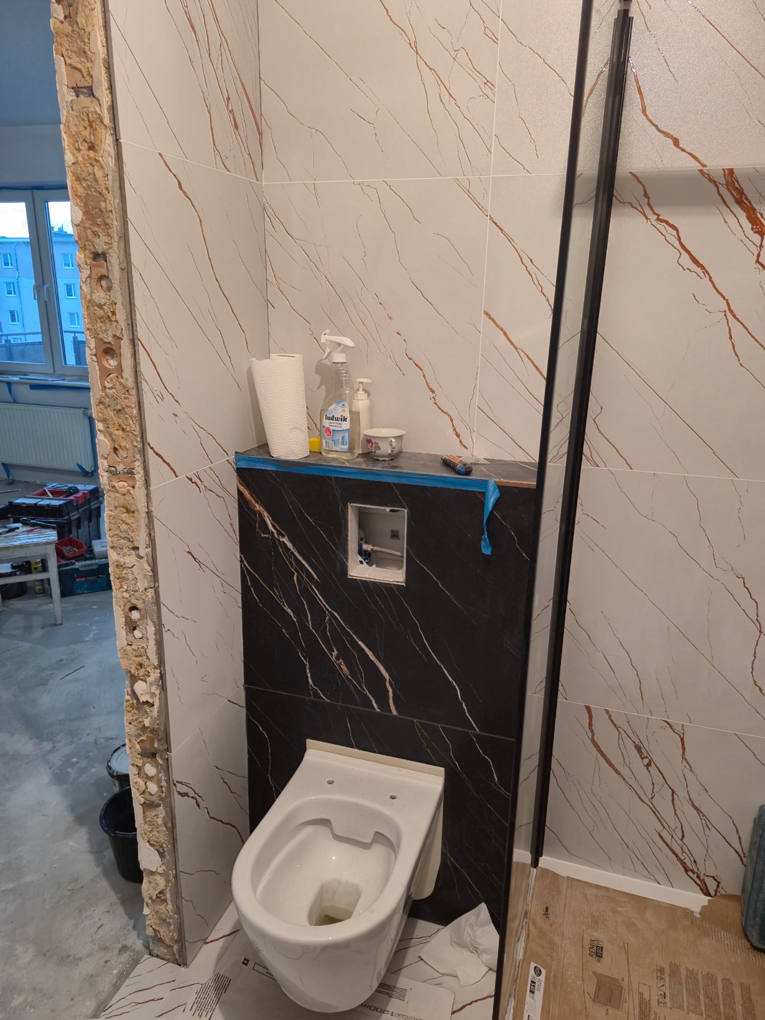 Niedokończona toaleta w łazience z białymi płytkami z brązowymi żyłkami i czarną zabudową geberitową, widoczne narzędzia i chemia gospodarcza, surowy stan wykończenia framugi drzwi.