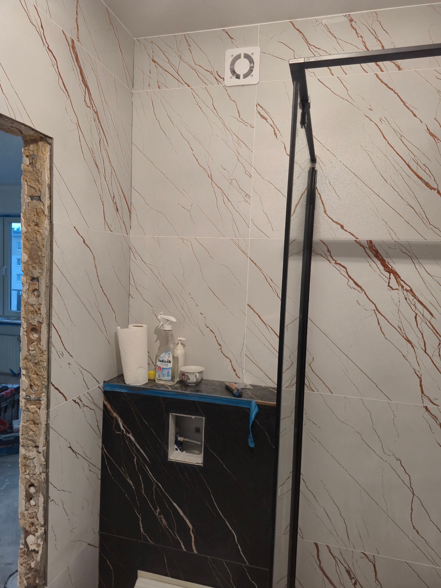 Wnętrze łazienki w trakcie prac wykończeniowych: częściowo położone płytki imitujące marmur z brązowymi żyłami, czarna zabudowa WC, widoczna konstrukcja kabiny prysznicowej, akcesoria i chemia...