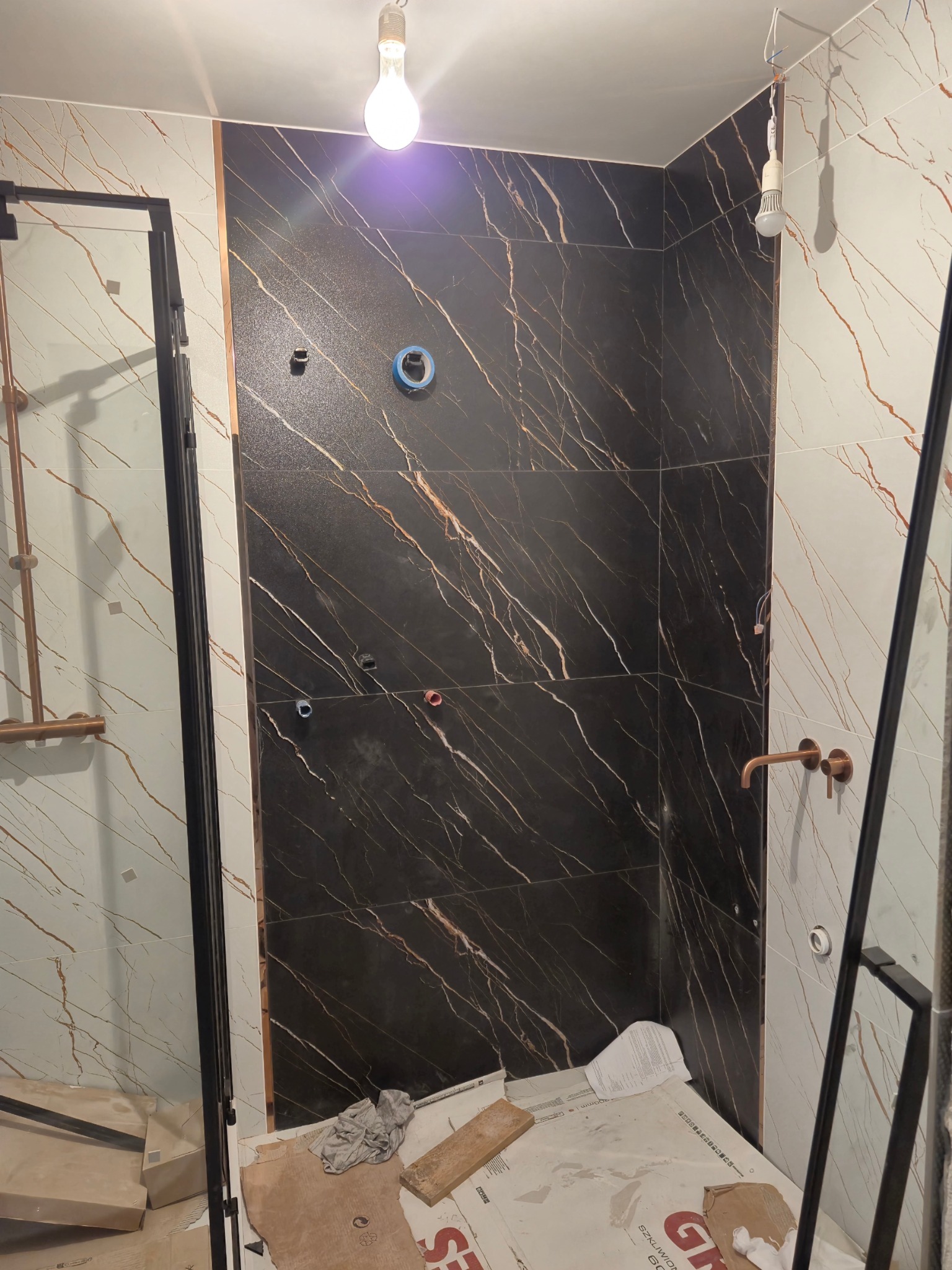 Łazienka w trakcie montażu płytek: ściana wyłożona czarnymi płytkami z białymi żyłami, obok ściana z białych płytek z brązowymi żyłami, widoczne otwory na instalacje, na podłodze zabezpieczona...