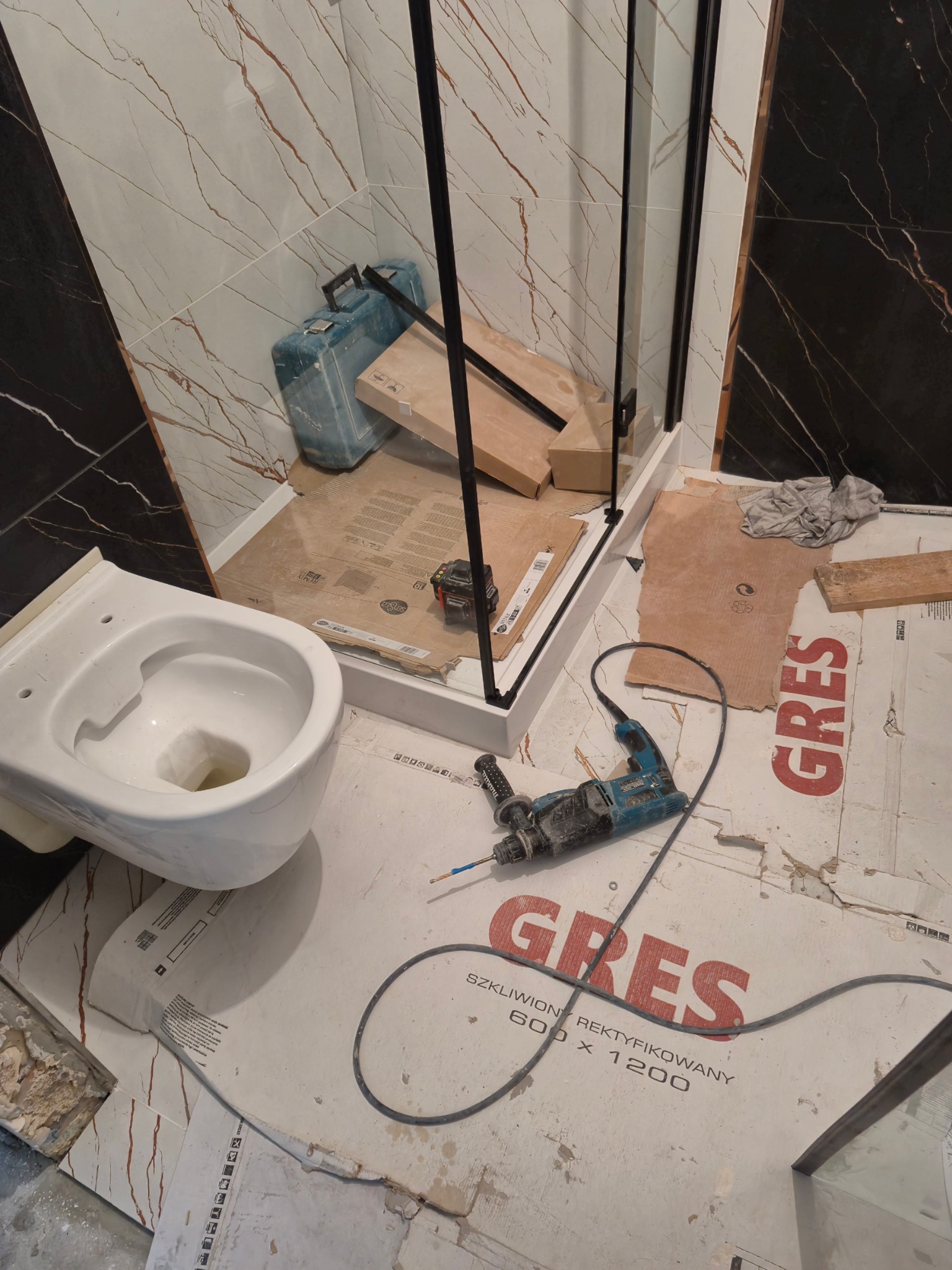 Niezamontowana muszla klozetowa, częściowo zmontowana kabina prysznicowa z czarnymi profilami, wiertarka udarowa oraz skrzynka narzędziowa na podłodze wyłożonej kartonem ochronnym z napisem 'GRES'...